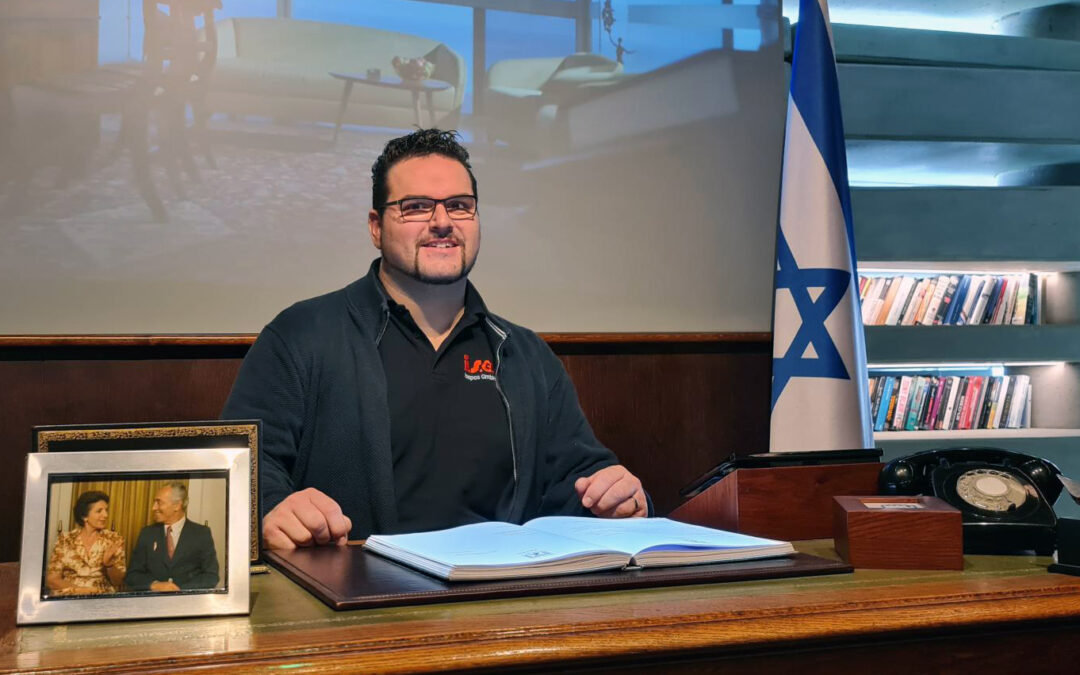 Geschäftsführer Martin Singer sitzt an einem großen Schreibtisch. Vor ihm ein großes Buch, hinter ihm die Fahne von Israel.